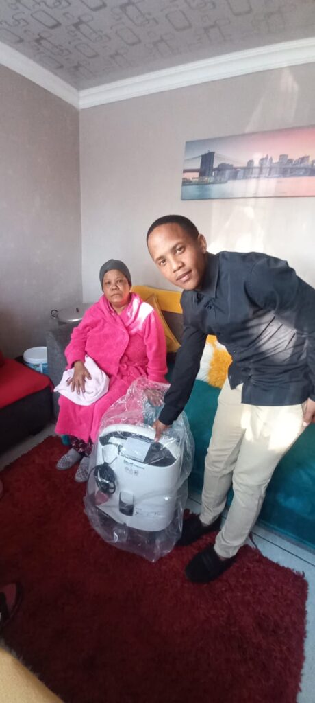 Man in a dark shirt presents a wrapped washing machine in plastic to an elderly woman in a pink robe seated on a sofa at home.